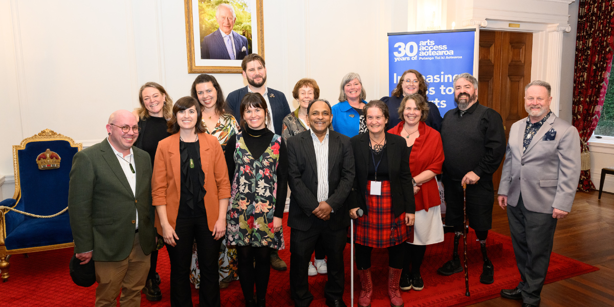 Arts Access Aotearoa staff group together for a staff photo at Government House. They stand in front of a blue pull-up banner which reads '30 Years of Arts Access Aotearoa. Increasing access to the arts'.